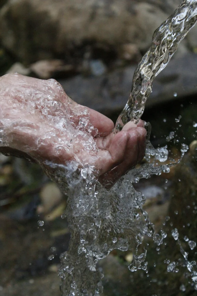 A close-up photograph capturing a pair of hands held together to catch clear, fast-flowing water, likely from a natural stream. The background is slightly blurred, emphasizing the clarity and motion of the water, evoking themes of purity, sustainability, and the vital importance of clean water resources.