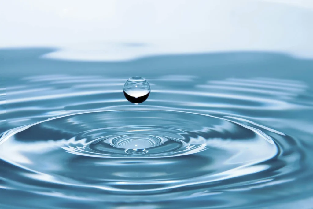  high-resolution macro photograph capturing a single water droplet suspended mid-air just before it touches the calm surface of a body of water, forming concentric ripples. The image highlights the purity, clarity, and fluid motion of water, symbolizing balance, conservation, and the precious nature of this life-sustaining resource. The soft blue tones convey serenity and cleanliness, making this image ideal for use in topics related to water conservation, environmental sustainability, hydration, natural resources, and the global water crisis. Perfect for eco-friendly blogs, water-saving campaigns, and wellness or science-related websites.