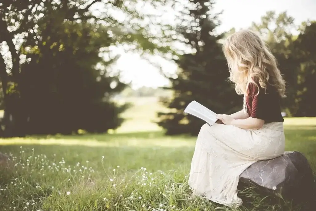The Art of Slow Reading breathes softly in this serene image of a young woman lost in a book while sitting on a stone in a sunlit meadow. Surrounded by tall trees and delicate wildflowers, the scene exudes tranquility and mindfulness. A celebration of disconnecting from digital noise and reconnecting with nature and literature, this visual is perfect for websites and blogs focused on wellness, self-care, book recommendations, and slow living.